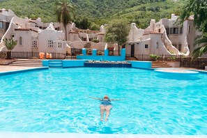 Una piscina con una persona flotando, rodeada por un edificio blanco de techos de tejas y un paisaje montañoso.