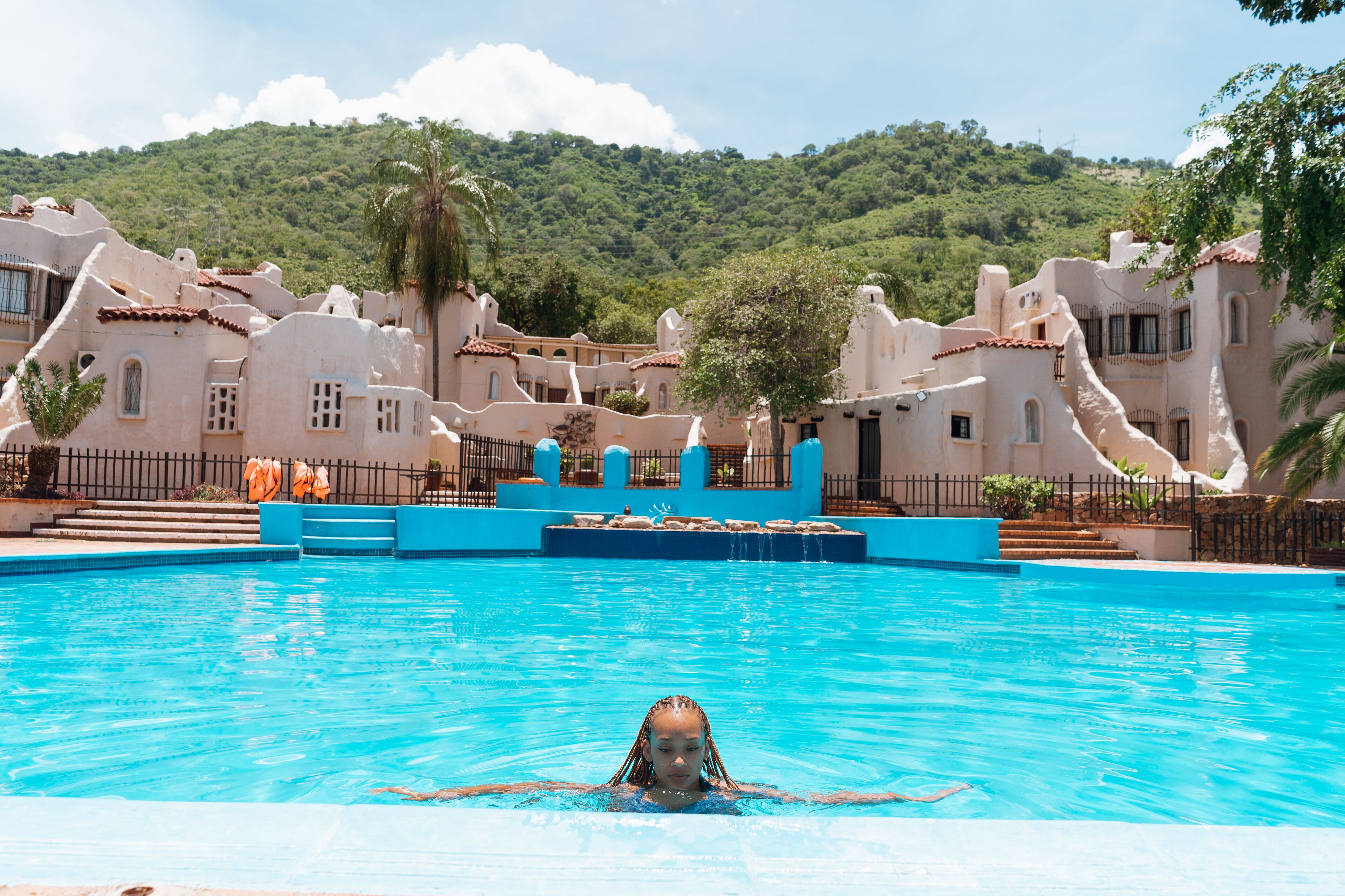 A swimming pool with a person in the water, surrounded by buildings with unique architecture and a hilly landscape in the background.