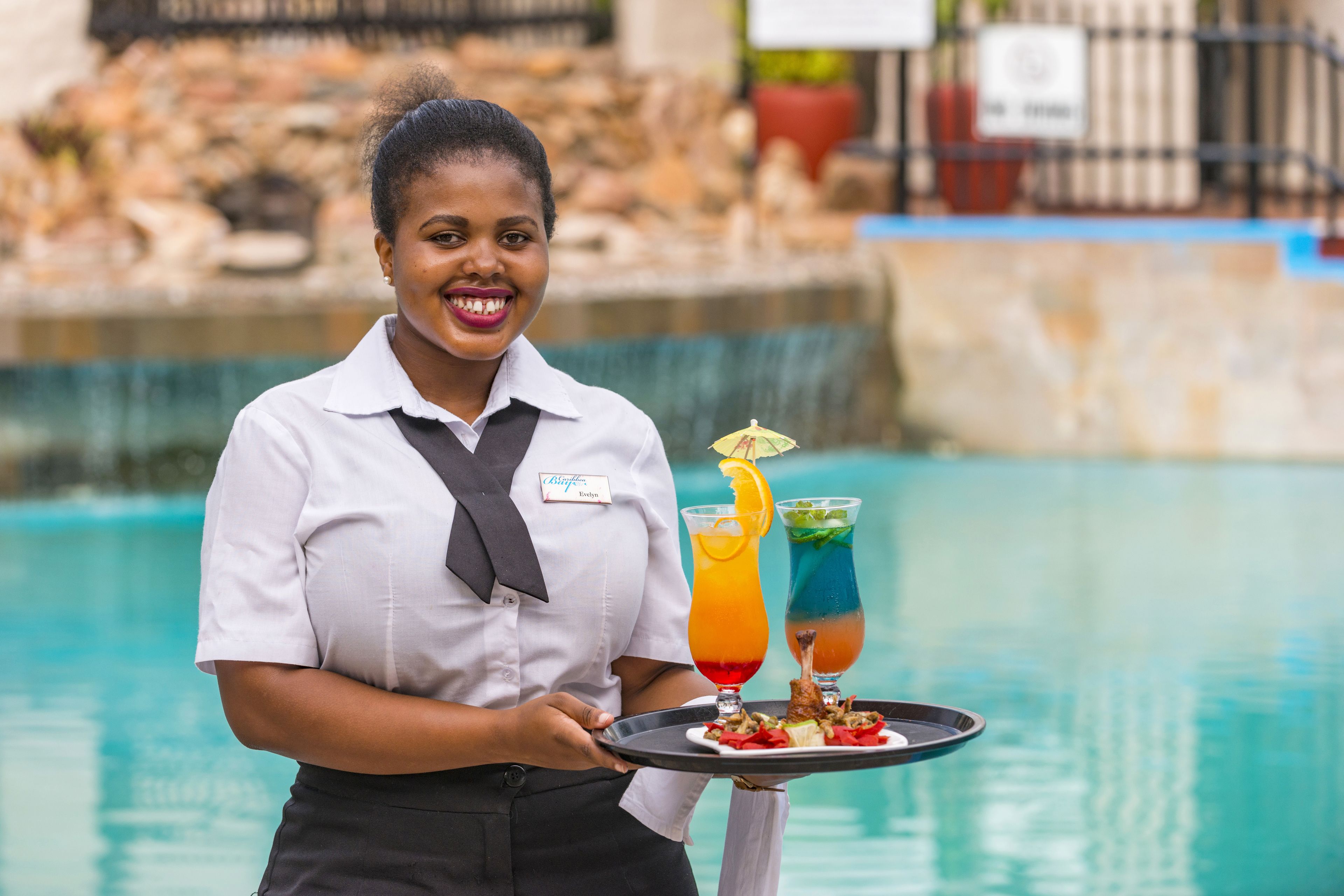 A person holding a tray with two drinks and a plate of food near a pool.