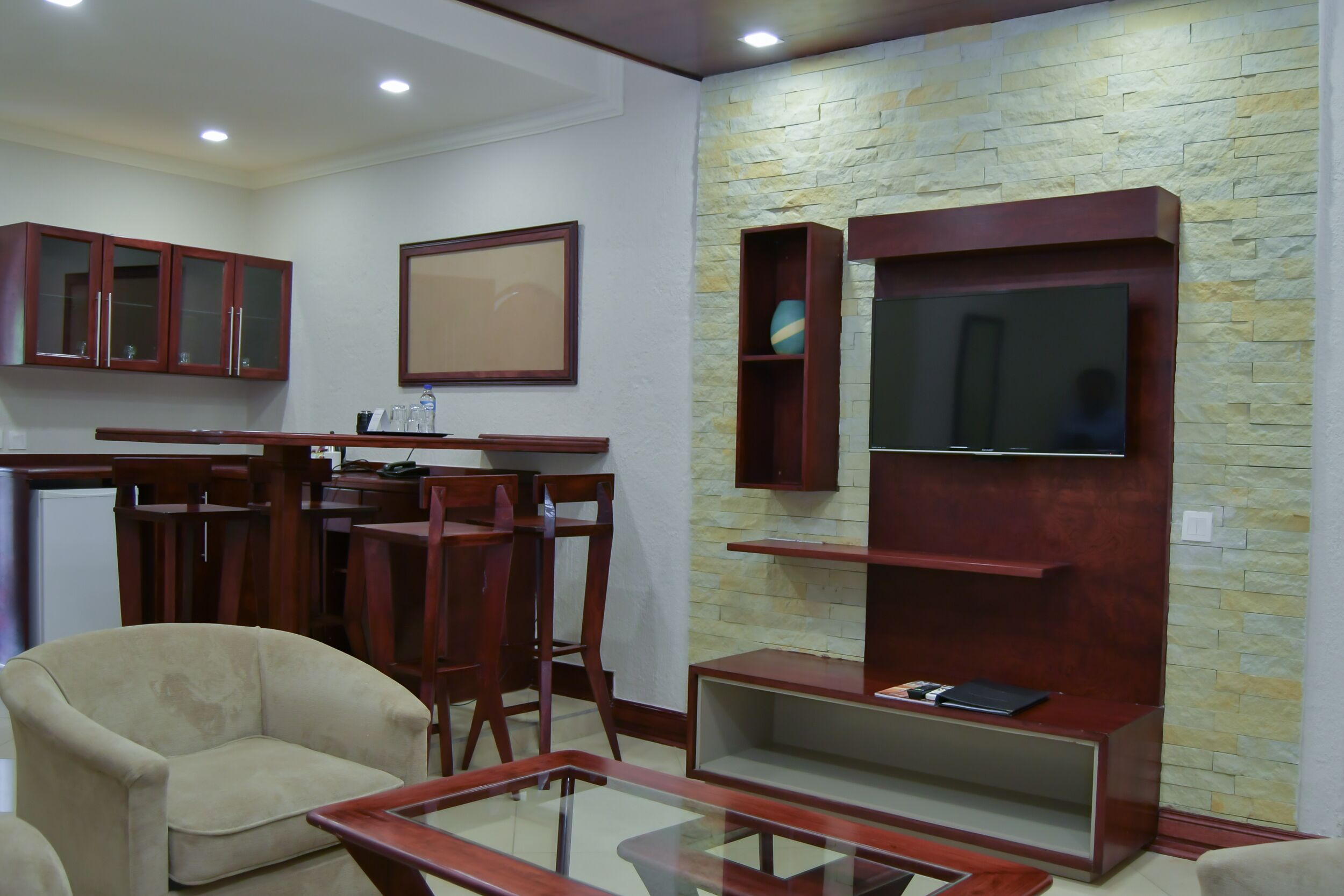 Una sala de estar con sofá, sillas, una mesita de centro de vidrio, armarios de madera, televisor y pared de piedra.
