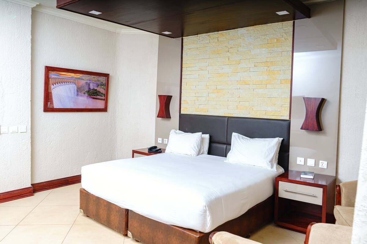 Quarto de hotel com uma cama grande, cabeceira de madeira, duas mesas de cabeceira, um criado-mudo e uma moldura com um quadro na parede, além de uma parede com padrão de tijolos.