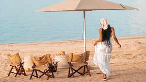 En strandscene med en kvinde i en hvid kjole, der går væk fra et bord med en beige parasol, to strandstole og en flettet kurv.