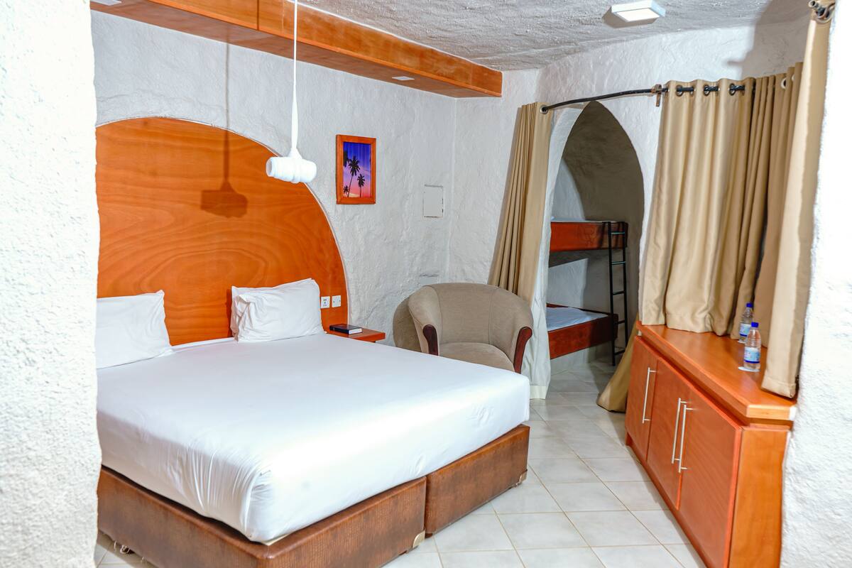 Quarto de hotel com uma cama grande, cabeceira de madeira, uma mesinha, uma cadeira e um guarda-roupa.