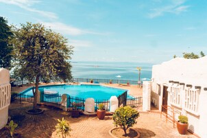 Zona de piscina con vista al mar, rodeada de plantas en macetas y un edificio blanco.