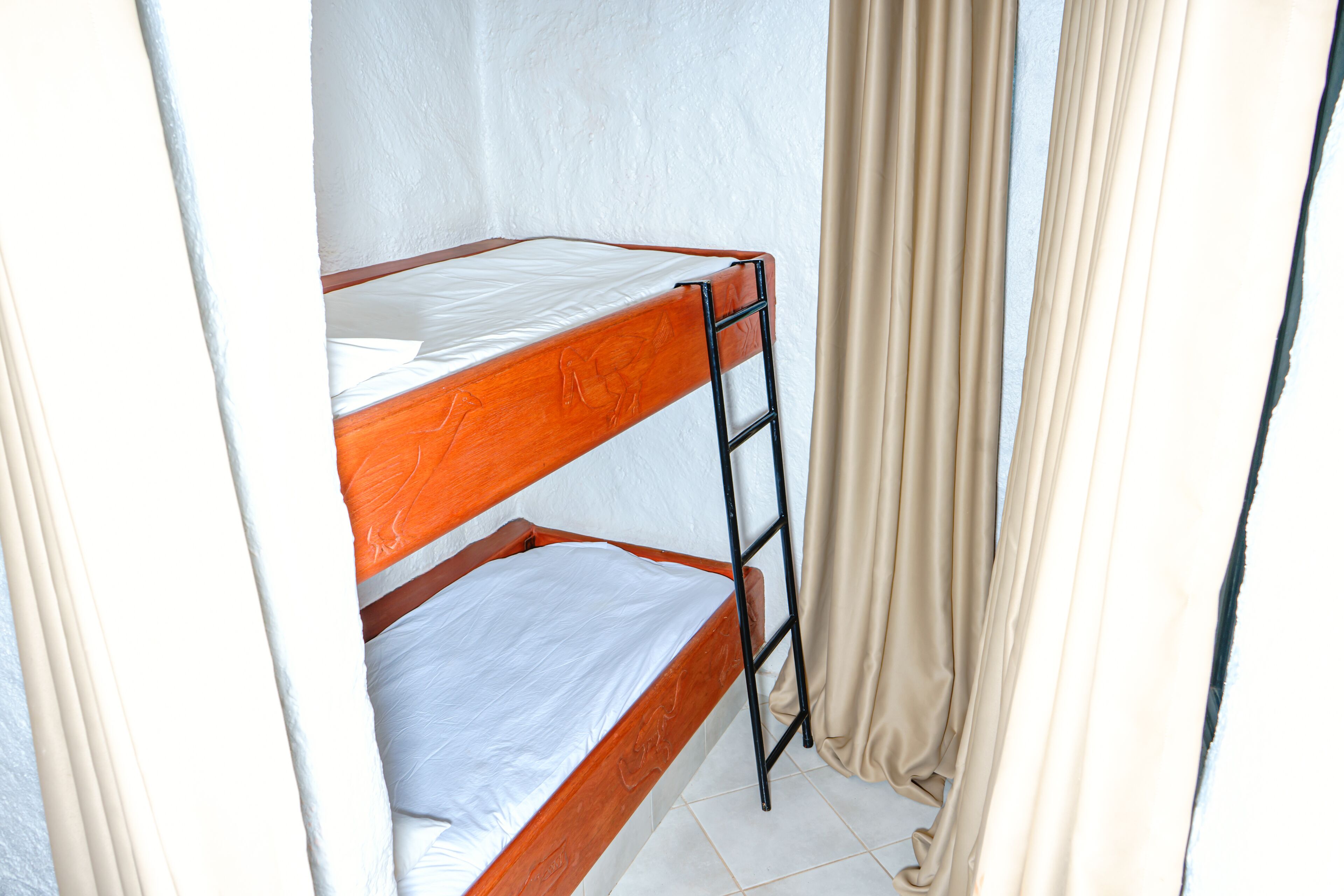 Una cama individual superpuesta con escalera, sábanas blancas y cortinas color beige.