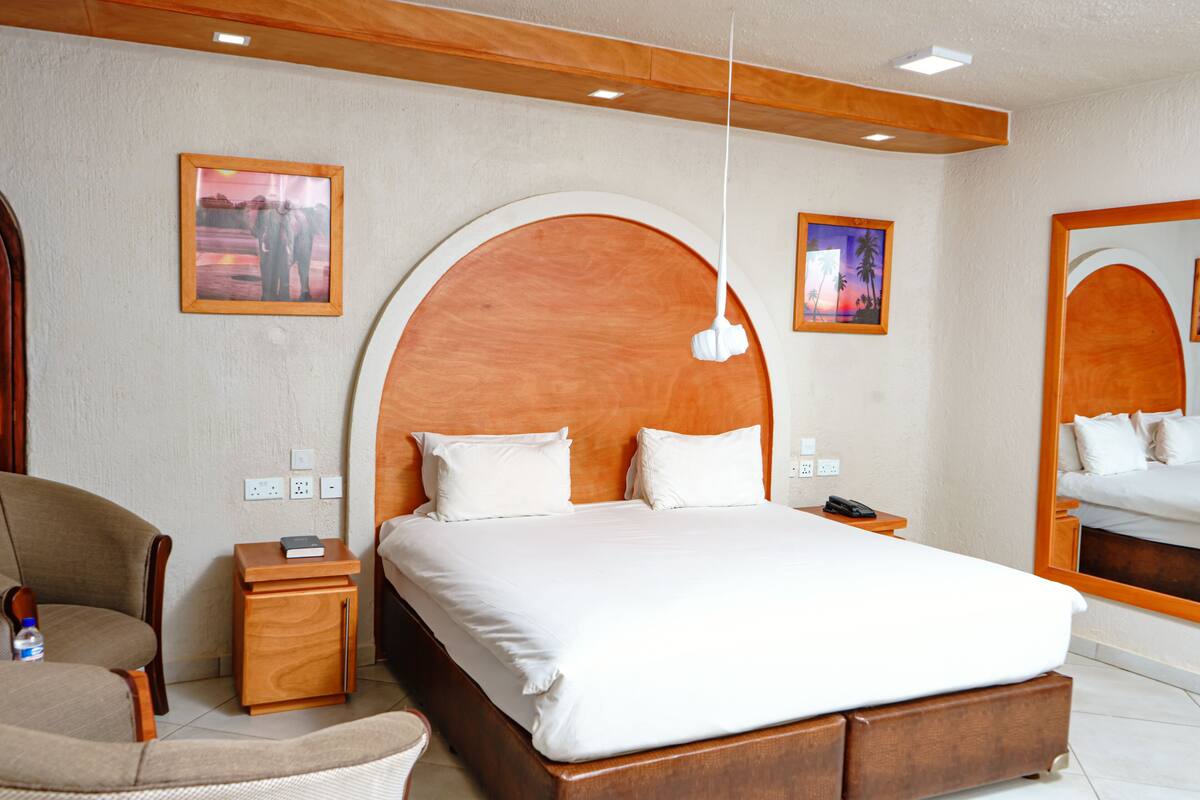 Quarto de hotel com uma cama grande, cabeceira de madeira, mesa de cabeceira com abajur, uma cadeira e duas fotos emolduradas na parede.