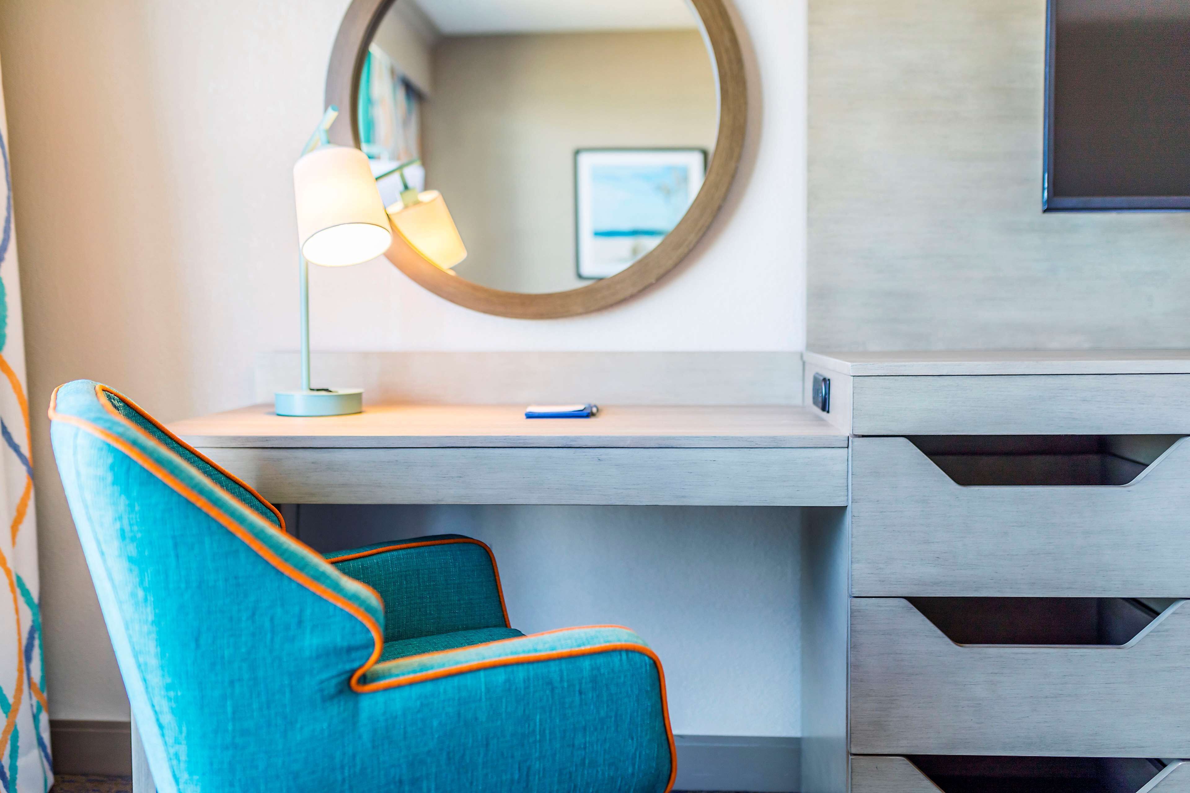 A modern room with a teal chair, a desk with drawers, a lamp, and a round mirror on the wall.