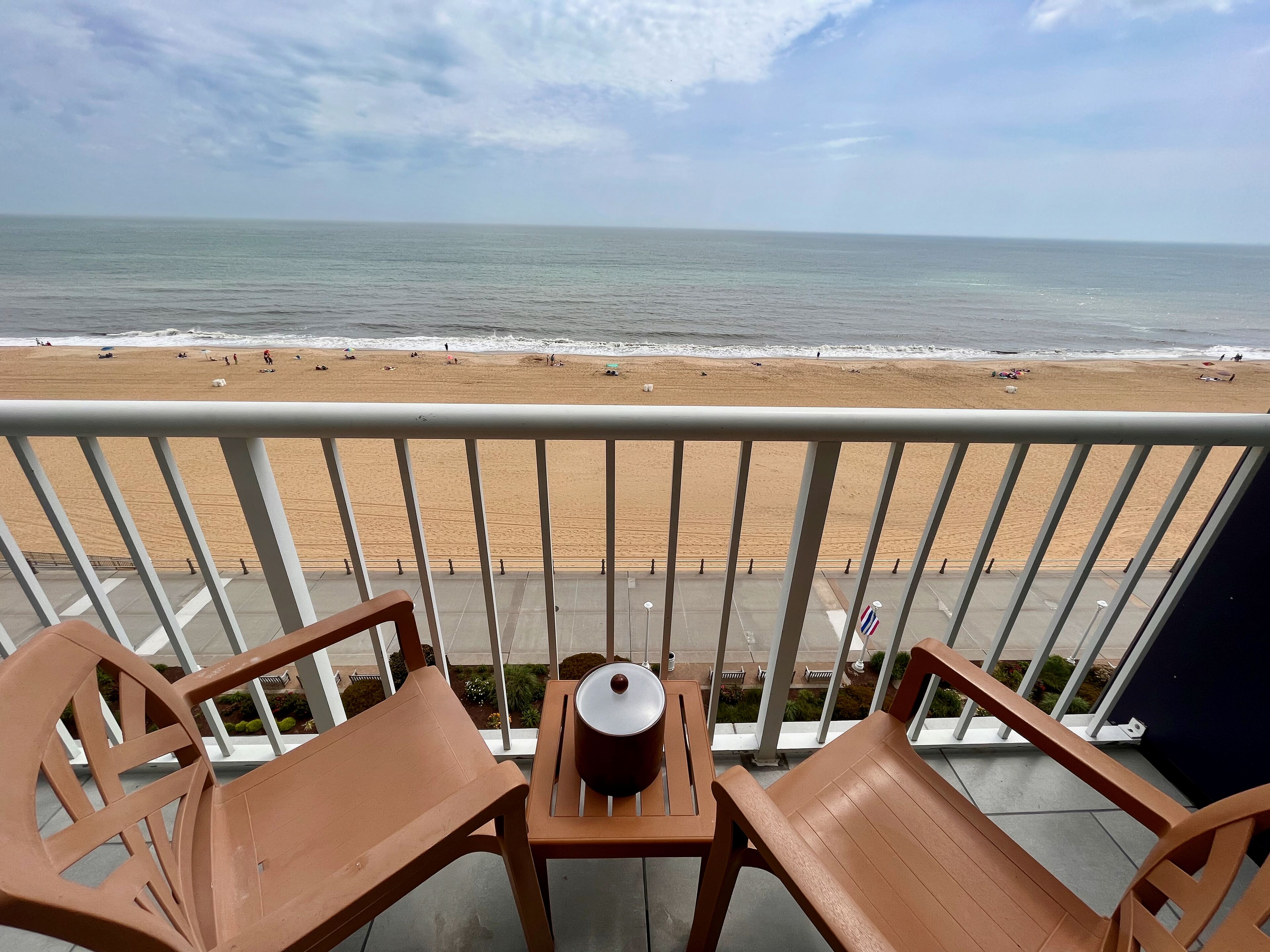 A balcony with two wooden chairs and a small table, overlooking a sandy beach and the ocean.