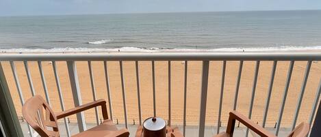 A balcony with two wooden chairs and a table, overlooking a beach with the ocean and a clear sky.