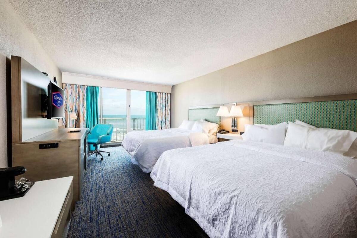 A hotel room with two beds, a desk, a TV, a chair, and a view of the ocean.
