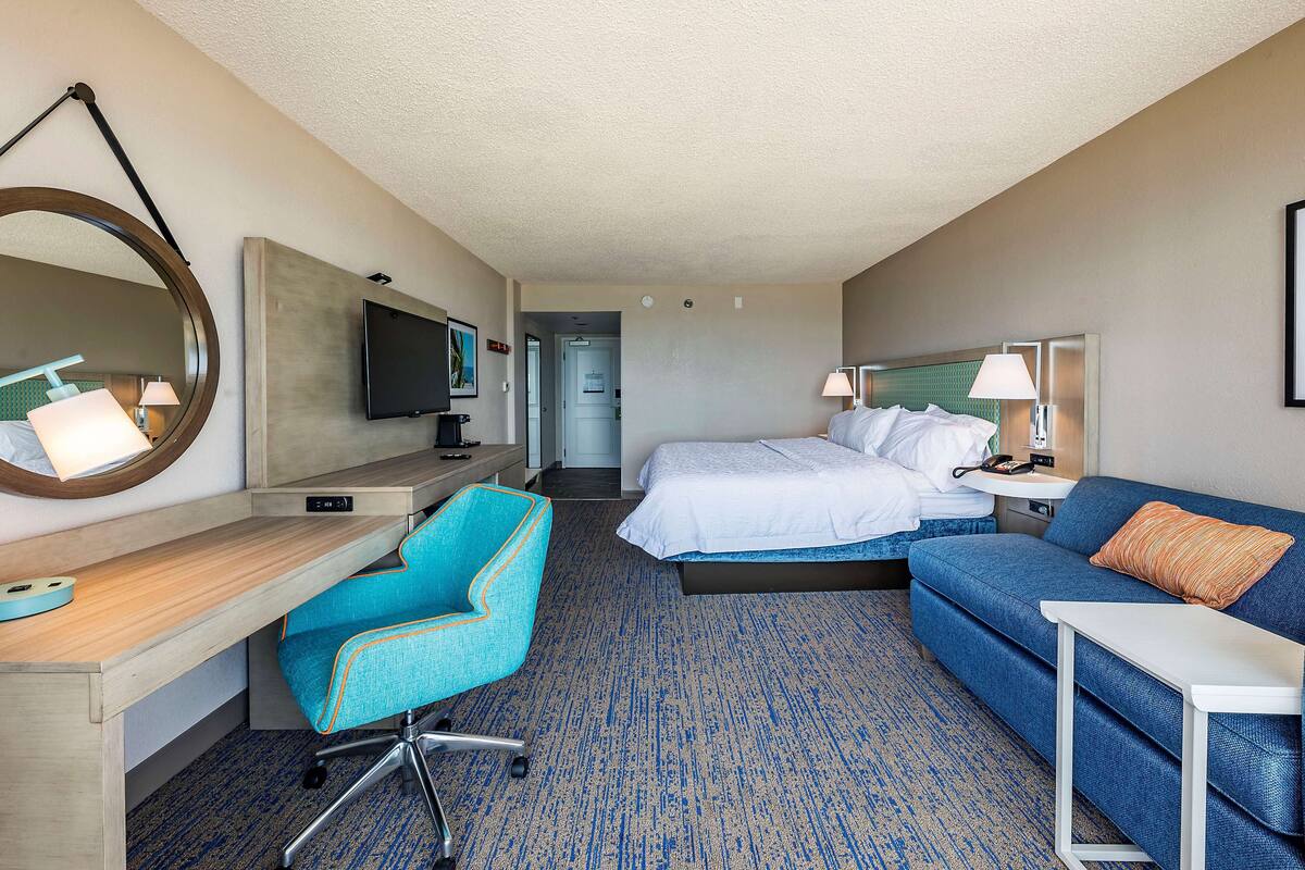 A hotel room with a desk, a blue armchair, a bed with white bedding, a blue sofa, and a television mounted on the wall.