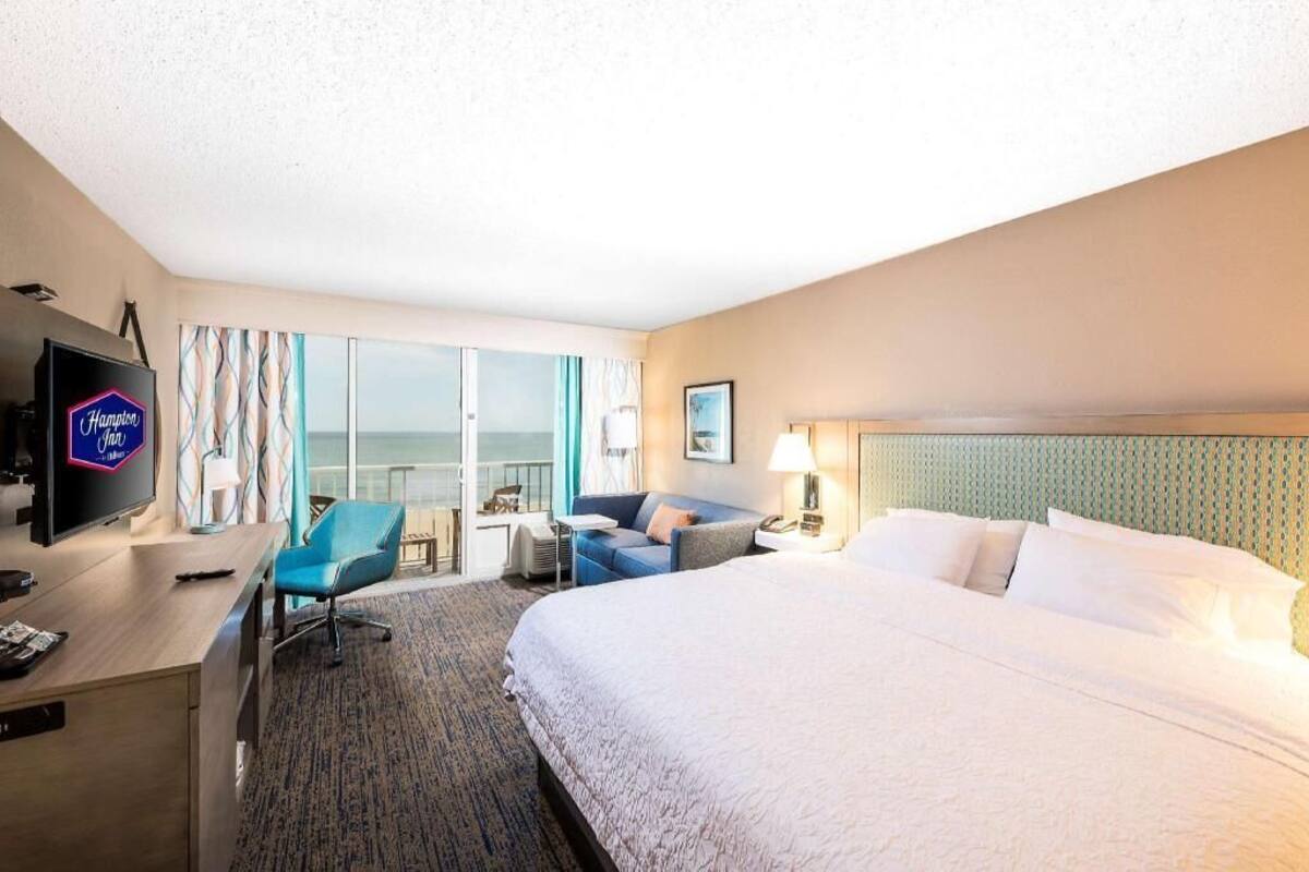 A hotel room with a bed, a desk with a television, a chair, a small table, and a view of the ocean through large windows.
