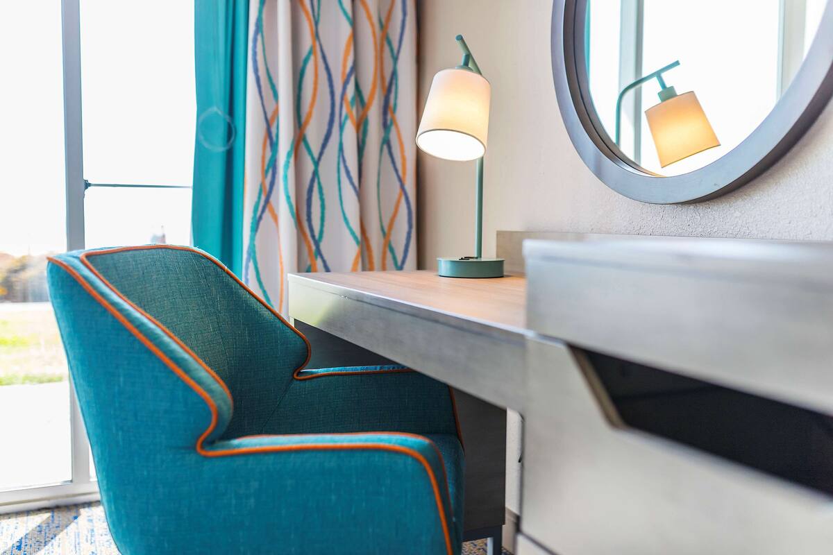 A teal upholstered chair with orange trim, a round mirror, and a table lamp with a white shade.