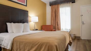 Quarto de hotel com uma cama grande, mesa de cabeceira, abajur, cadeira, janela com cortinas e porta.