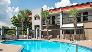 A swimming pool with a metal ladder, surrounded by a concrete deck. There are chairs and tables under a covered area, and palm trees are visible. - Quality Inn & Suites Aiken East (Aiken)
