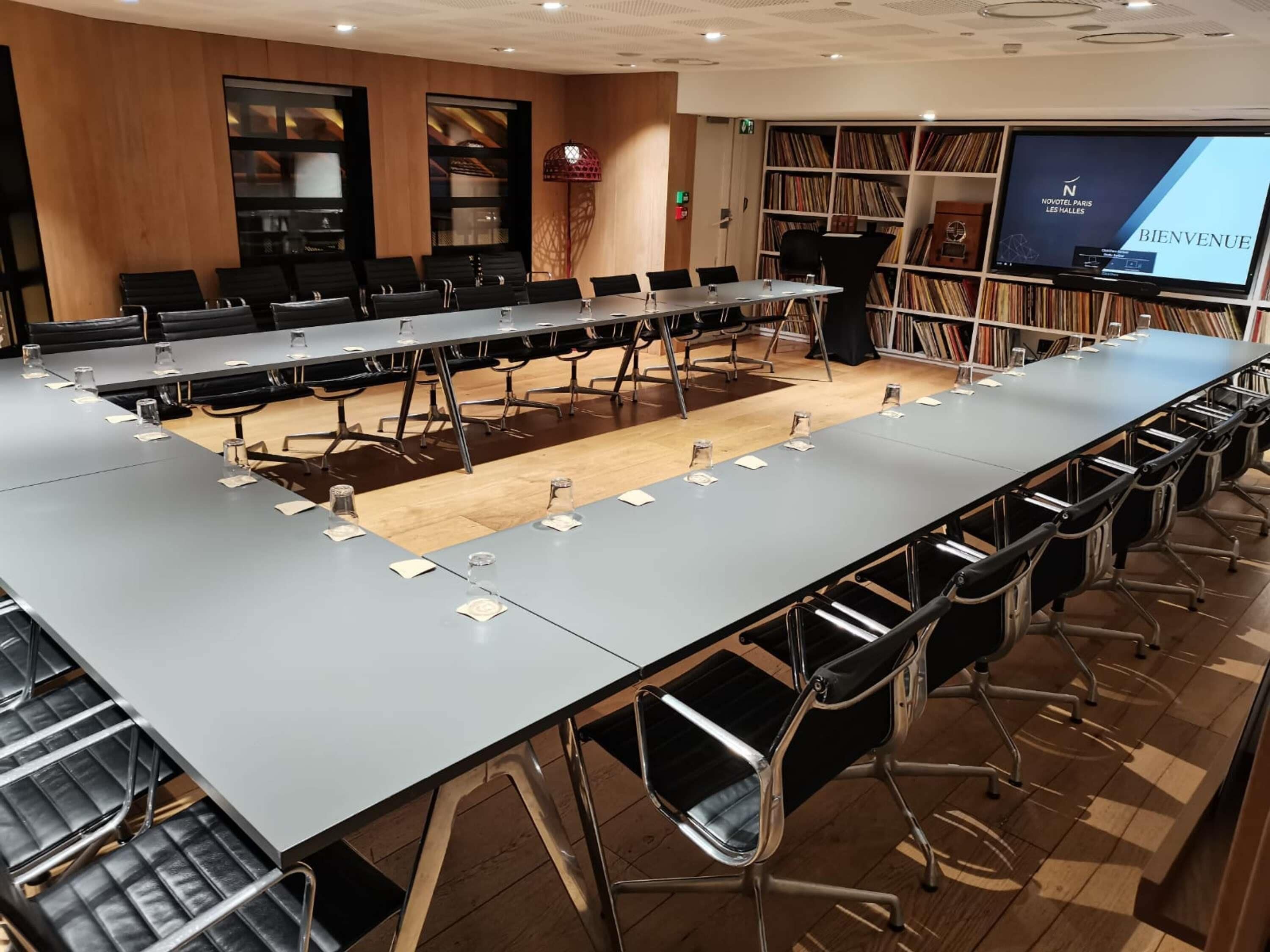 A conference room with a long table, chairs, a bookshelf, and a screen displaying 'BIENVENUE'.