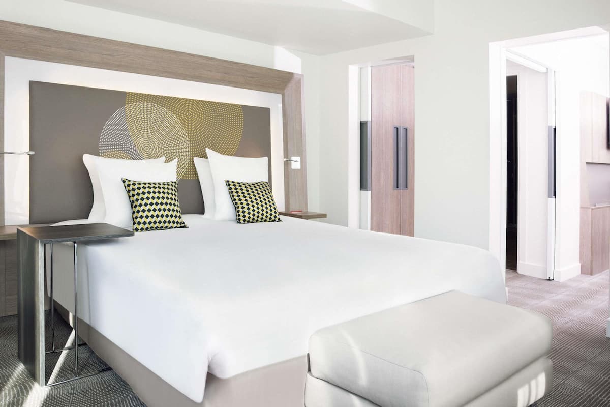A modern hotel room with a large bed, a bedside table, a wardrobe, and a door leading to another room.