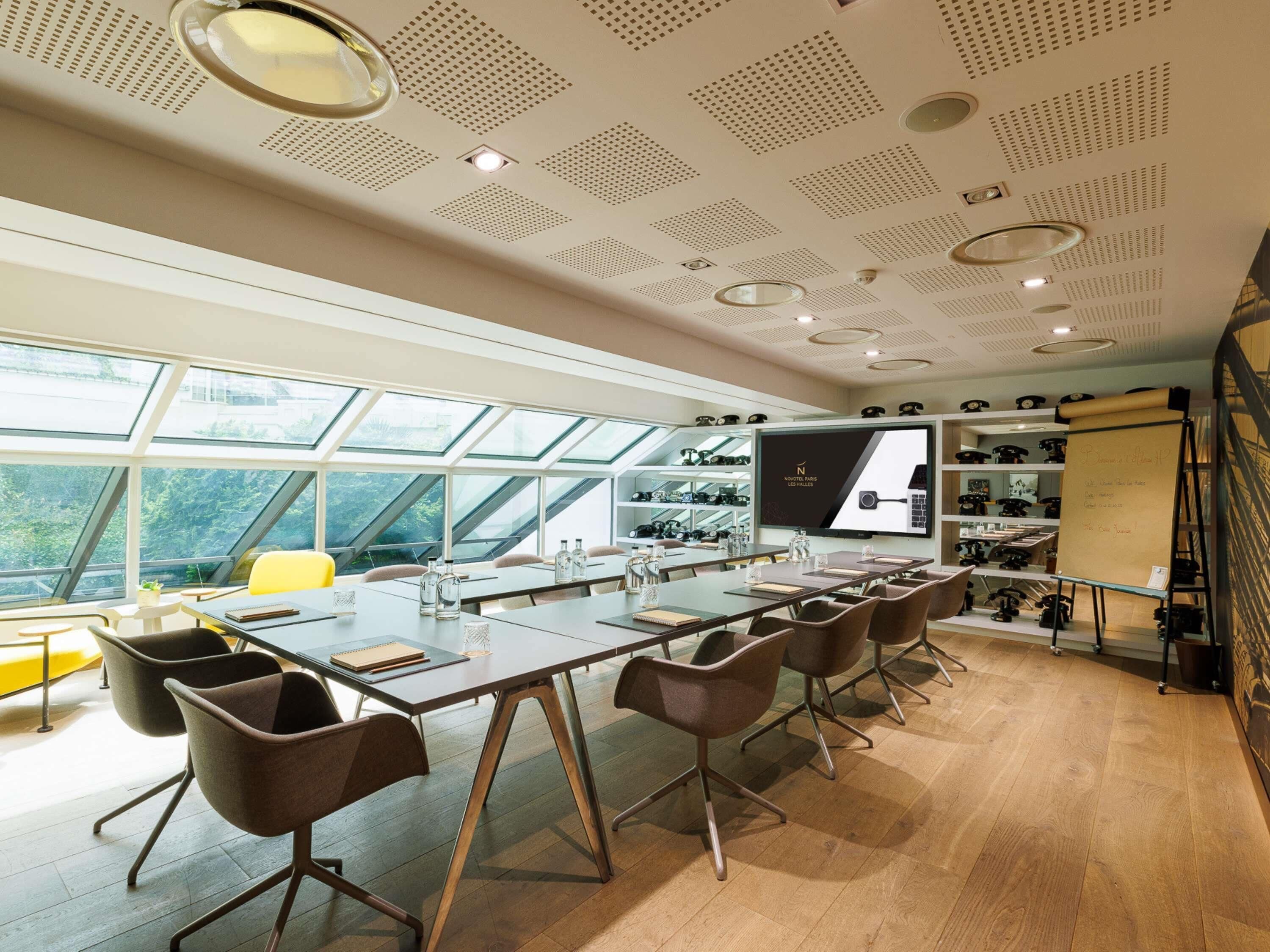 A modern conference room with a long table, brown chairs, a large screen, and a view of greenery through skylights.