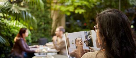 A person reading a magazine at an outdoor café with tables and chairs, surrounded by greenery.