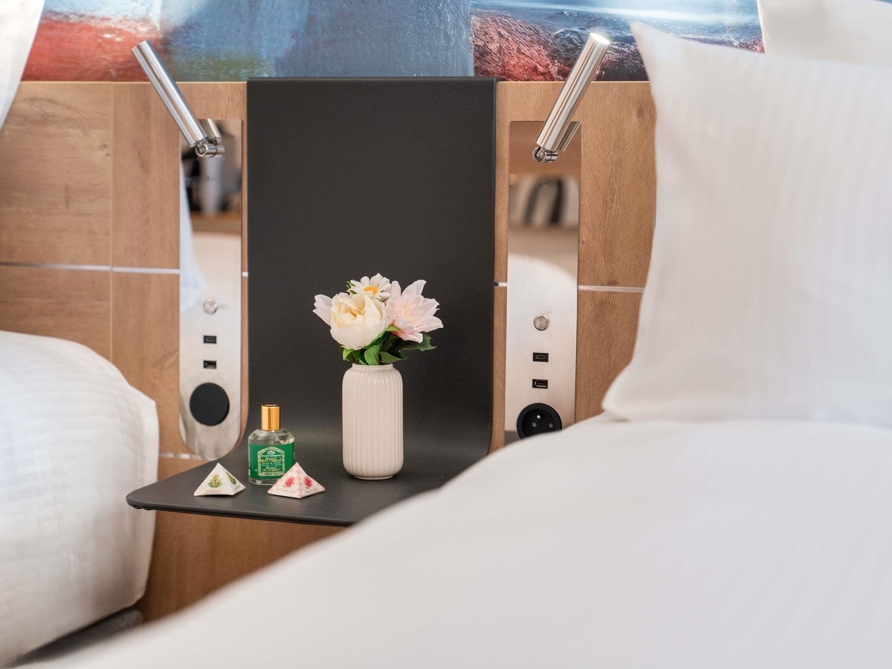 A hotel room with a bed, a small table with a vase of flowers, a bottle of perfume, and a mirror.