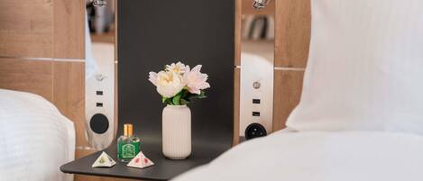 A hotel room with a bed, a small table with a vase of flowers, a bottle of perfume, and a mirror.