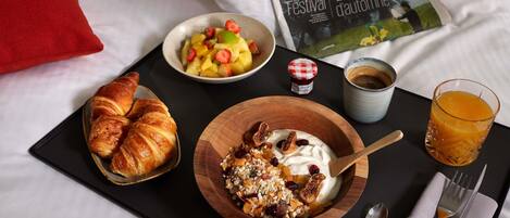 A breakfast tray with croissants, yogurt with nuts and dried fruits, a fruit bowl, a glass of orange juice, a coffee cup, and a magazine on a bed.
