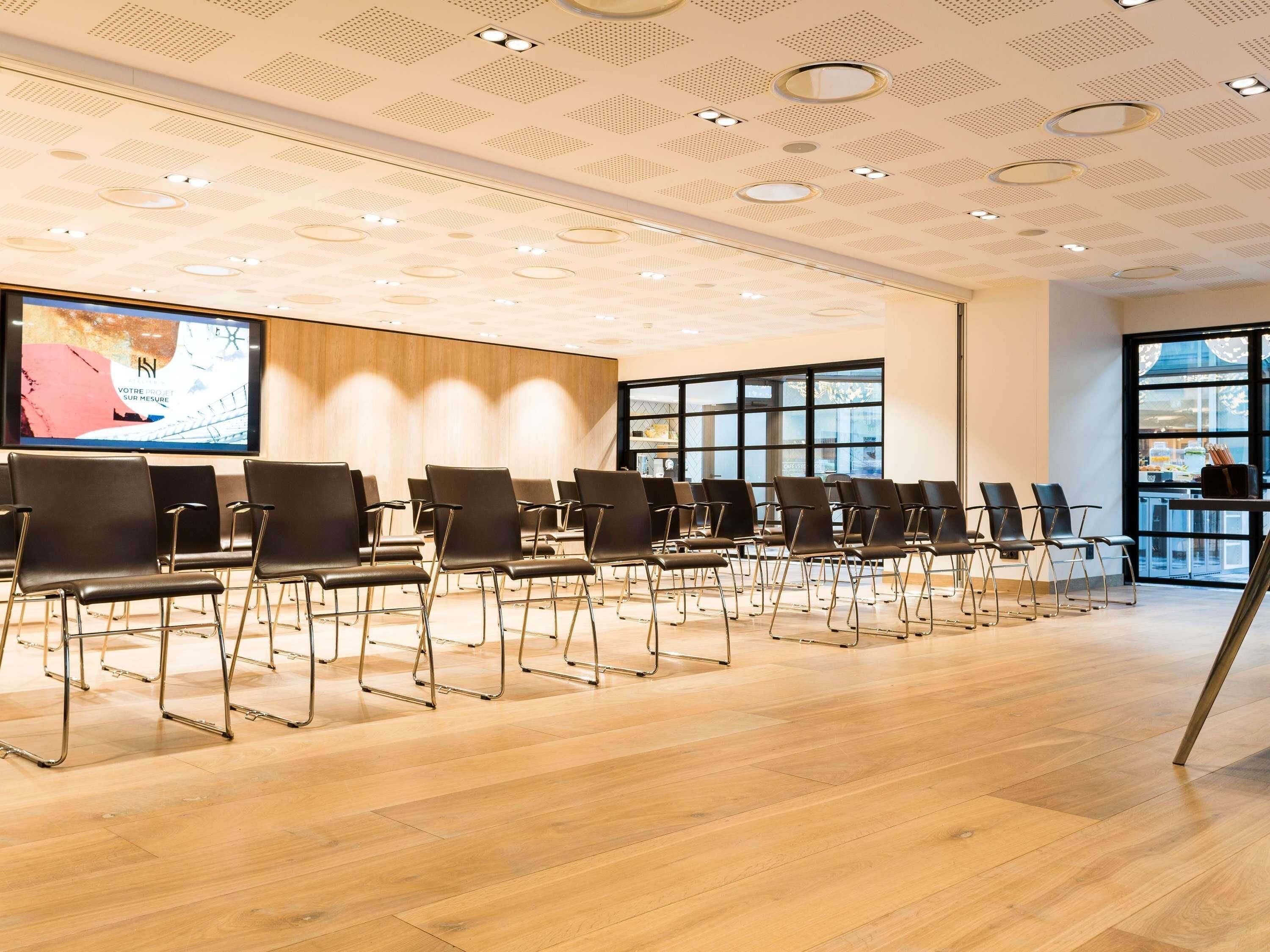 Un salón de conferencias con hileras de sillas negras, una pantalla grande y piso de madera.