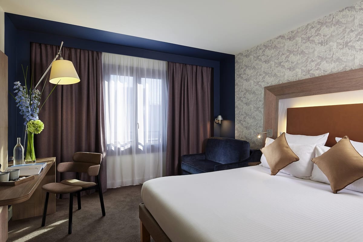 A hotel room with a bed, a desk, a chair, a lamp, and a window with curtains.