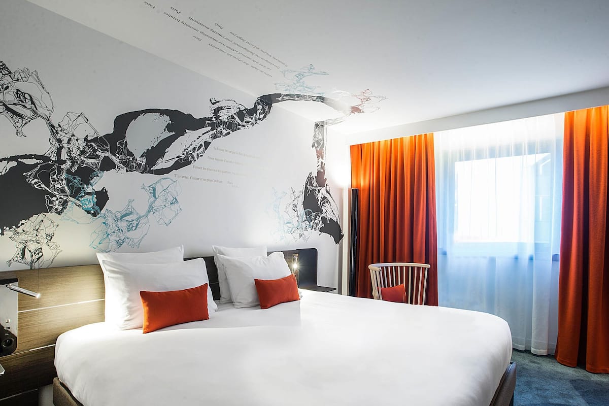 A hotel room with a large bed, a map mural on the wall, a chair, and a window with curtains.
