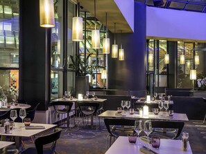 A modern restaurant interior with tables set for dining, pendant lighting, and large windows. - Novotel Paris Les Halles (Paris)