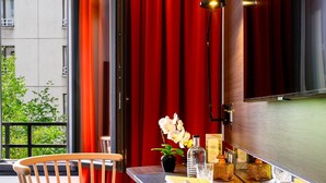 Una habitación moderna con una silla de madera, una mesita con un florero y un televisor, cortinas rojas.