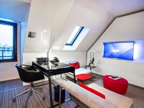 A modern, compact room with a desk, chair, sofa, and a small table with a plant. A large window provides natural light, and a TV is mounted on the wall.