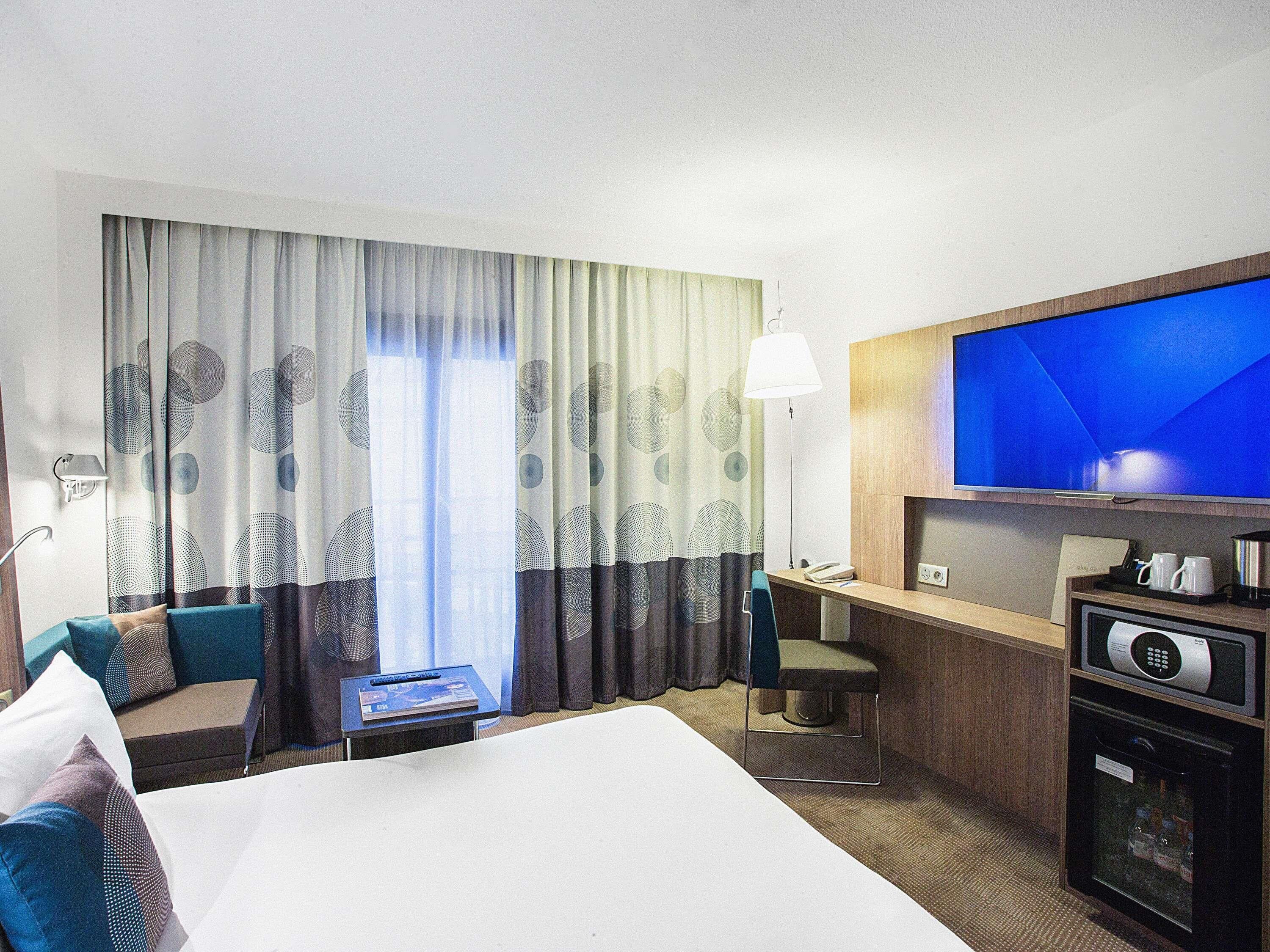 Habitación de hotel con cama, escritorio, silla, televisor, minibar y ventana con cortinas.