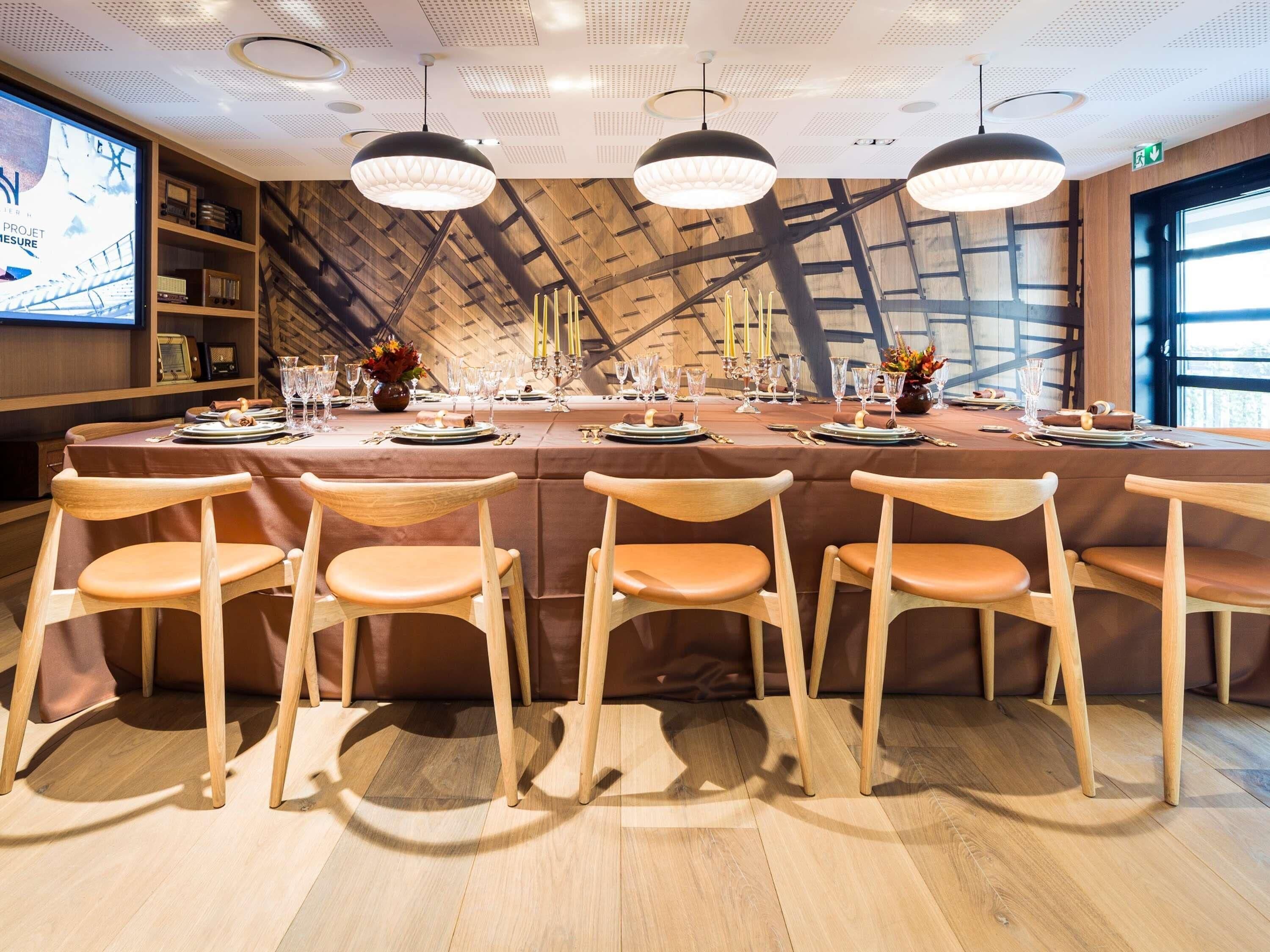 A dining area with a long table set for a meal, wooden chairs, pendant lights, and a wooden ceiling with a grid pattern.