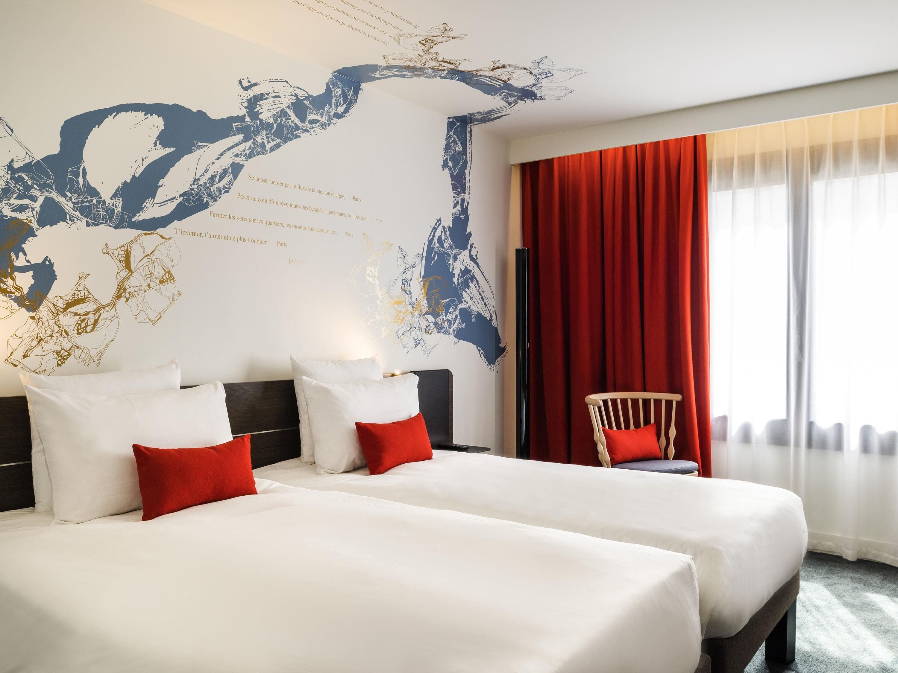 A hotel room with two beds, a red chair, a large abstract wall art, and a window with red curtains.