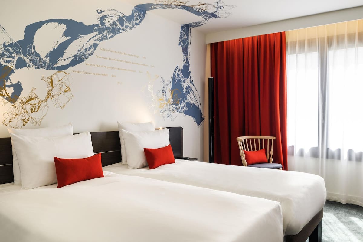 A hotel room with two beds, a red chair, a large abstract wall art, and a window with red curtains.
