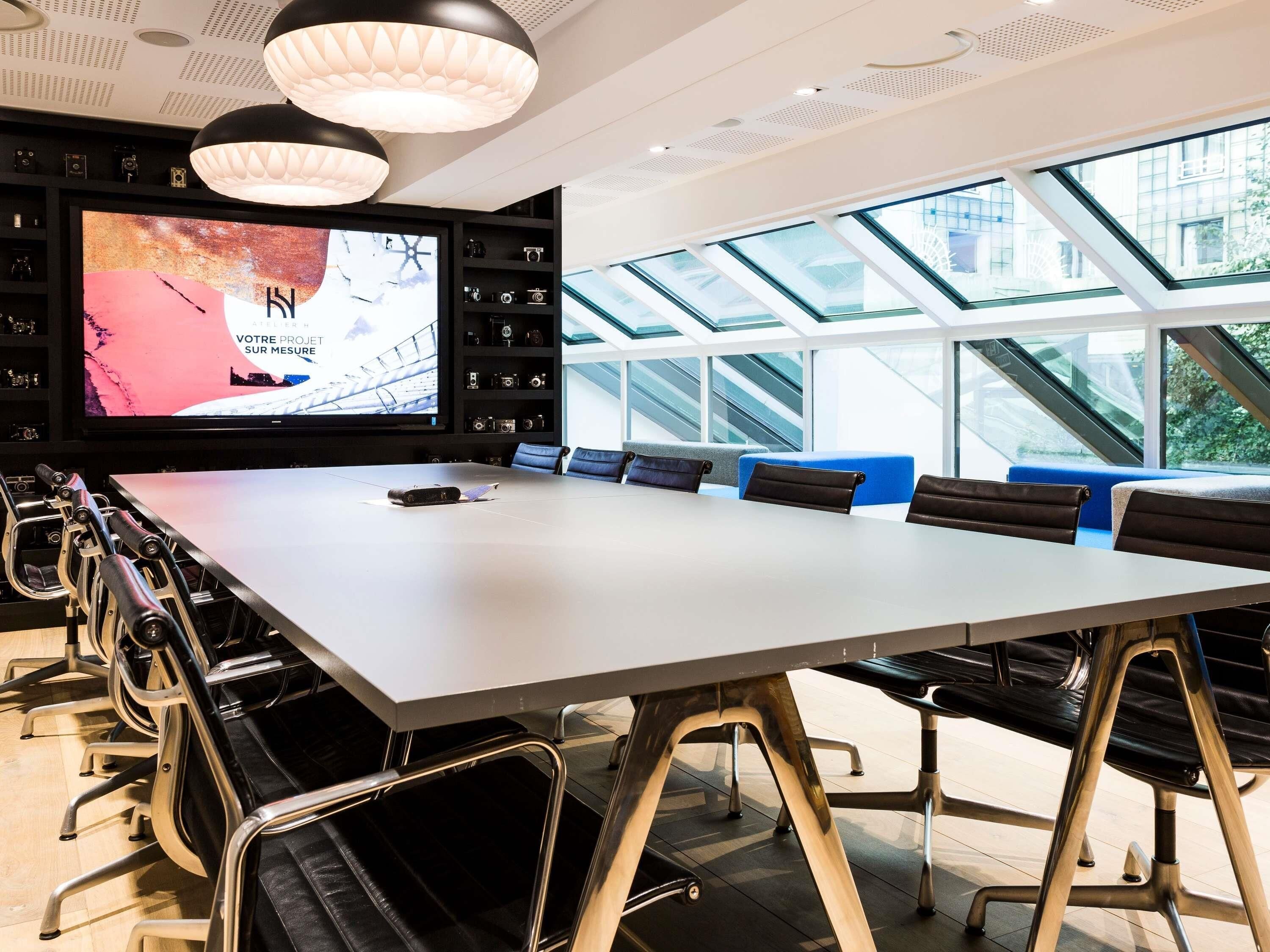 Uma sala de conferências com uma mesa comprida, cadeiras, uma tela grande e vista por claraboias.