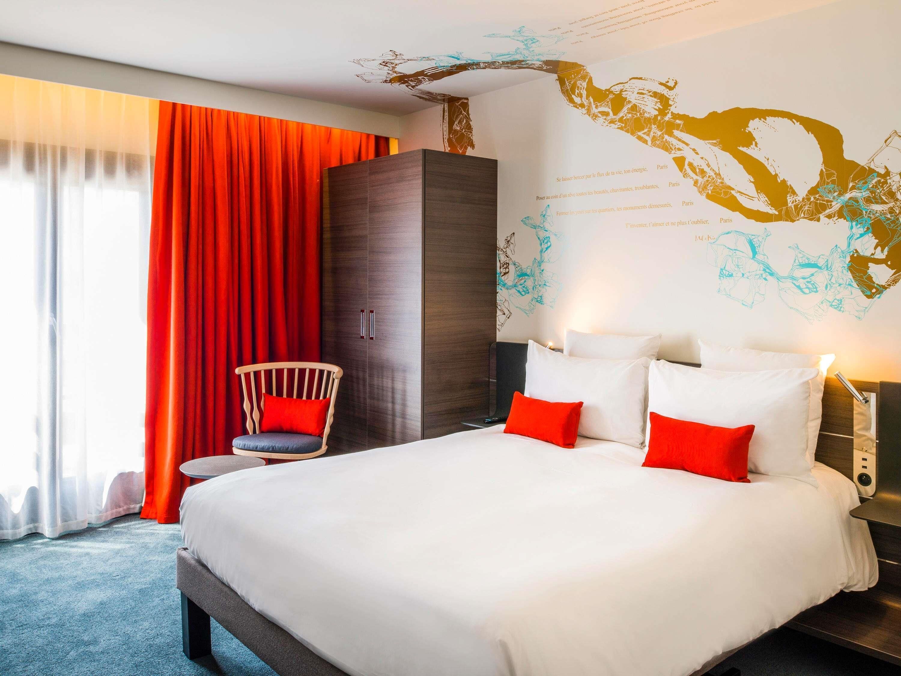 A hotel room with a large bed, red curtains, a wooden wardrobe, a chair, and a wall map.