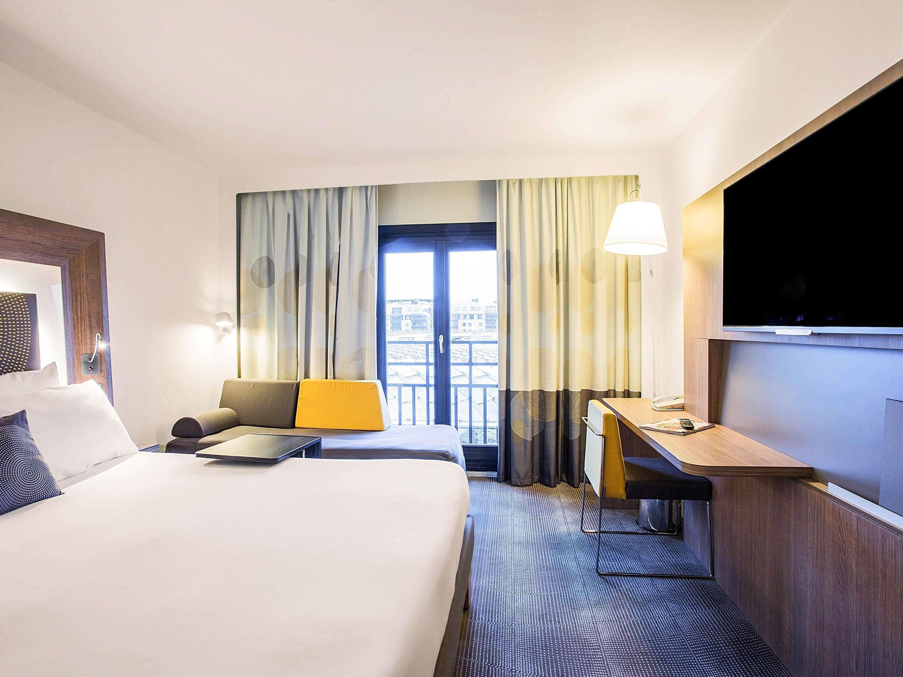 A hotel room with a bed, a desk, a chair, a TV, and a view of the city through the window.