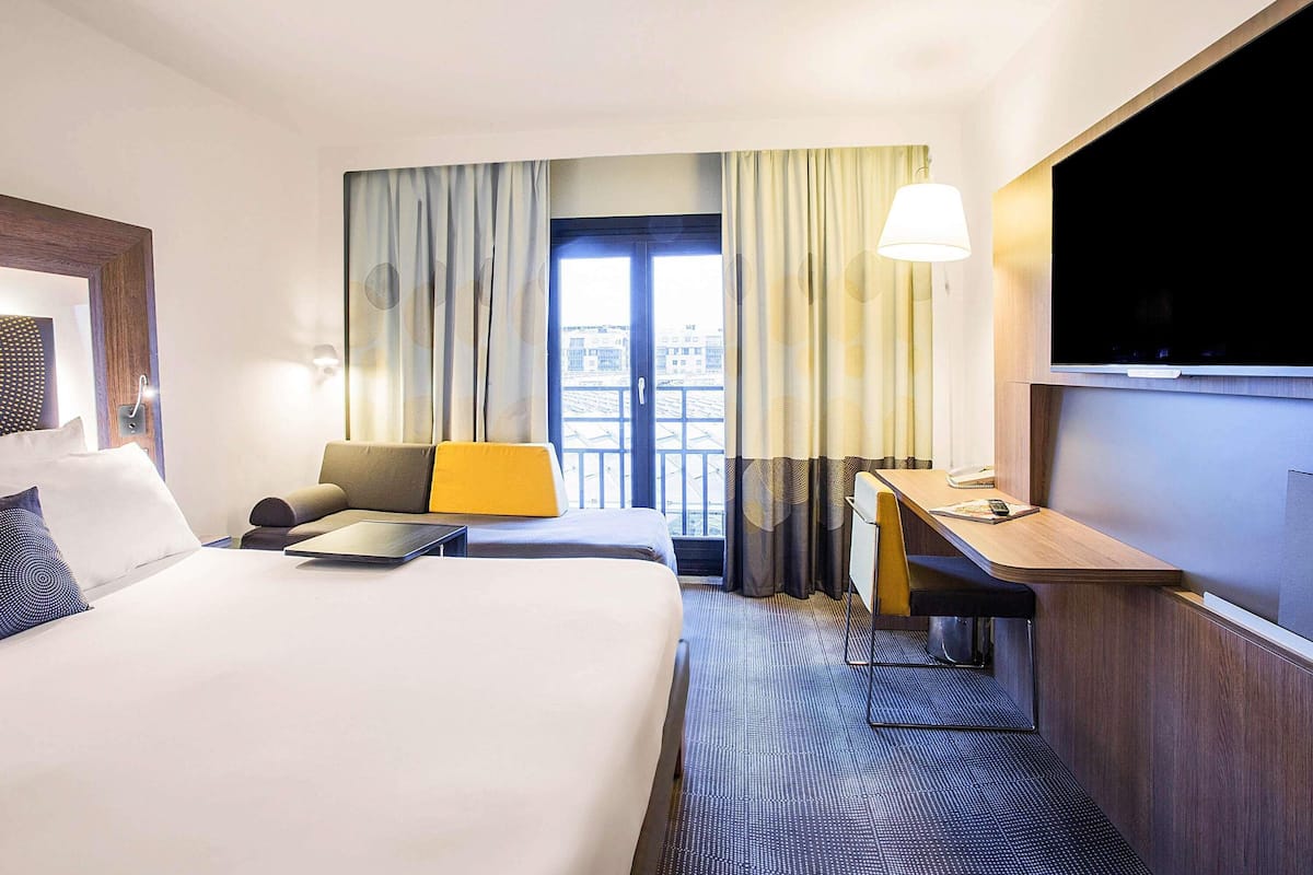 A hotel room with a bed, a desk, a chair, a TV, and a view of the city through the window.