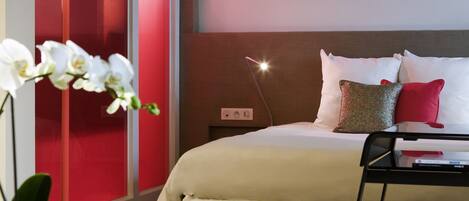 A modern hotel room with a bed, bedside table, lamp, and a potted plant.