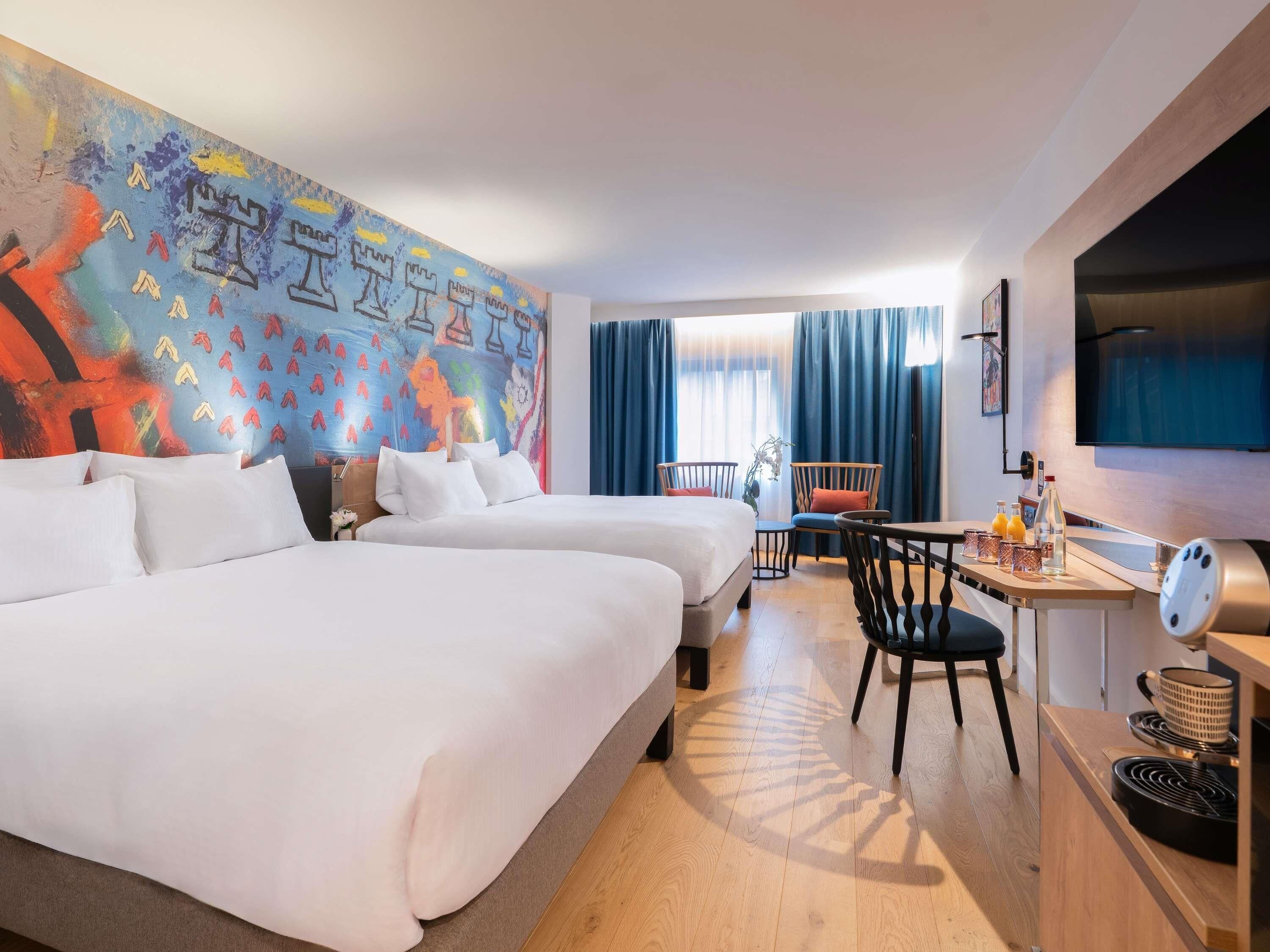 A hotel room with two beds, a desk with a chair, a TV, and a colorful mural on the wall.