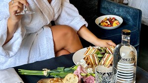 Una mujer con bata blanca sostiene una copa de vino, sentada a una mesa con un sándwich, una botella de Purezza y un bol de fruta.