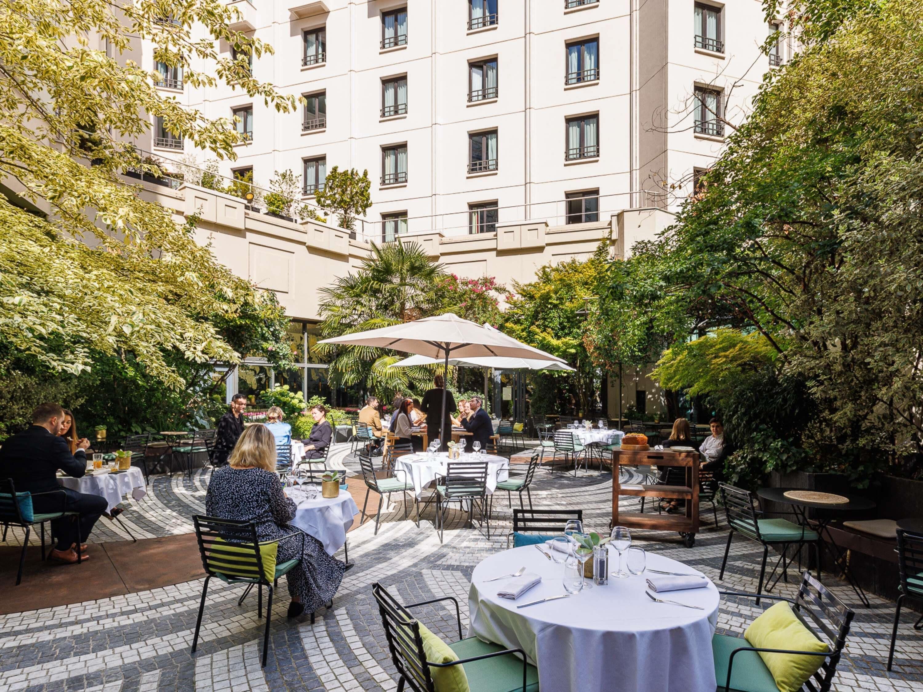 An outdoor dining area with tables and chairs, surrounded by greenery and a building in the background.