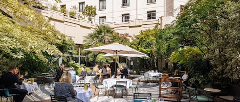 Un área de comedor al aire libre con mesas y sillas, rodeada de follaje y un edificio al fondo.