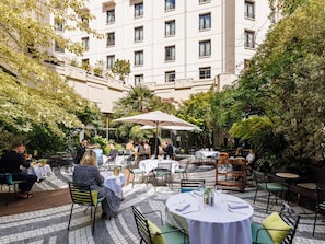 An outdoor dining area with tables and chairs, surrounded by greenery and a building in the background. - Novotel Paris Les Halles (Paris)