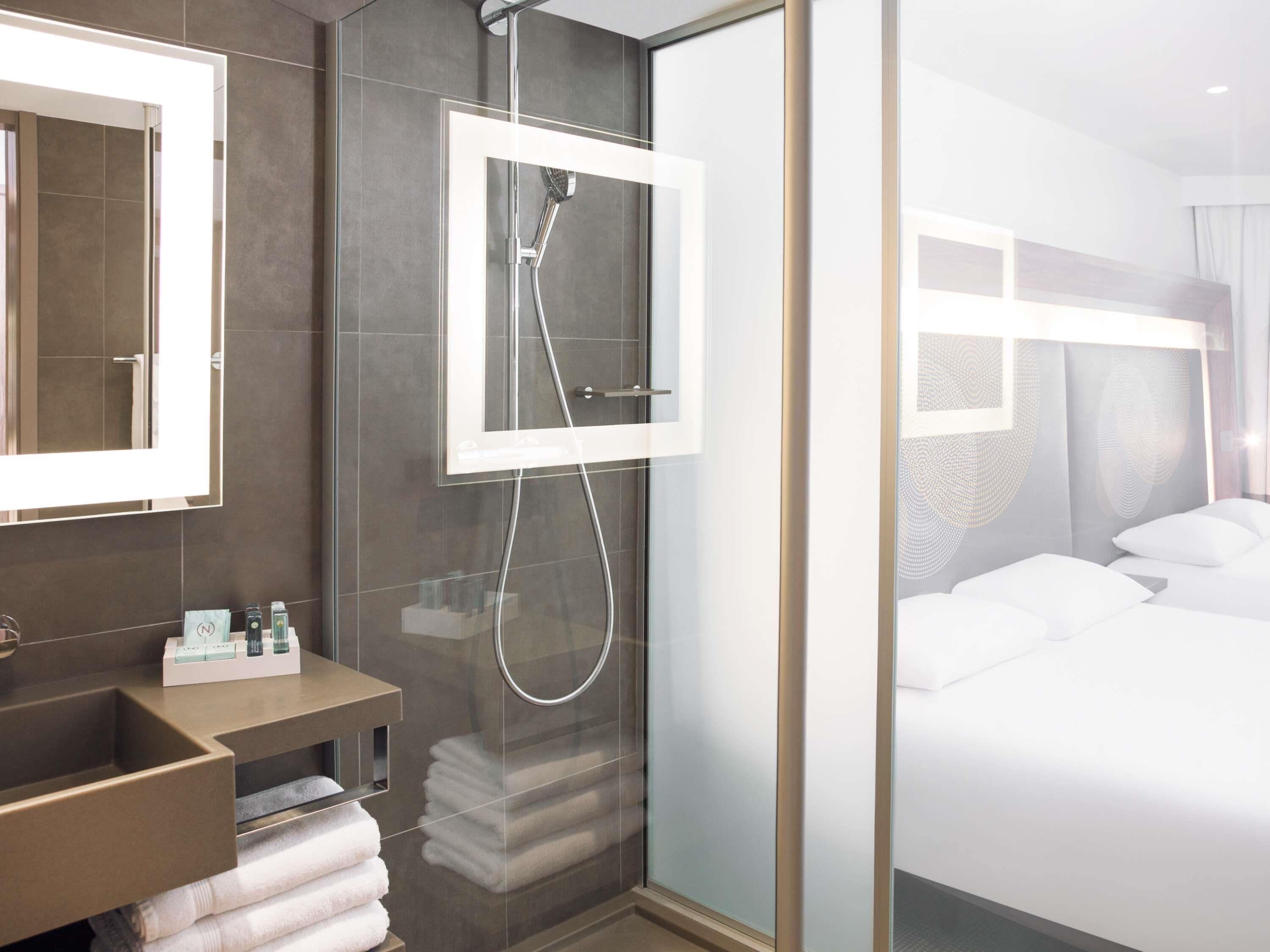 A modern hotel room with a glass-enclosed shower, a large mirror, a bedside table with toiletries, and a neatly made bed.