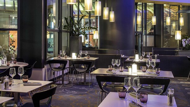 Interior de un restaurante moderno con mesas dispuestas para comer, iluminación ambiental y amplias ventanas.