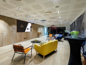 A modern hotel lobby with a wooden panel wall, a large screen displaying 'NOVOMATIC PARIS LES HALLES,' and a seating area with a yellow sofa and a wooden chair. - Novotel Paris Les Halles (Paris)