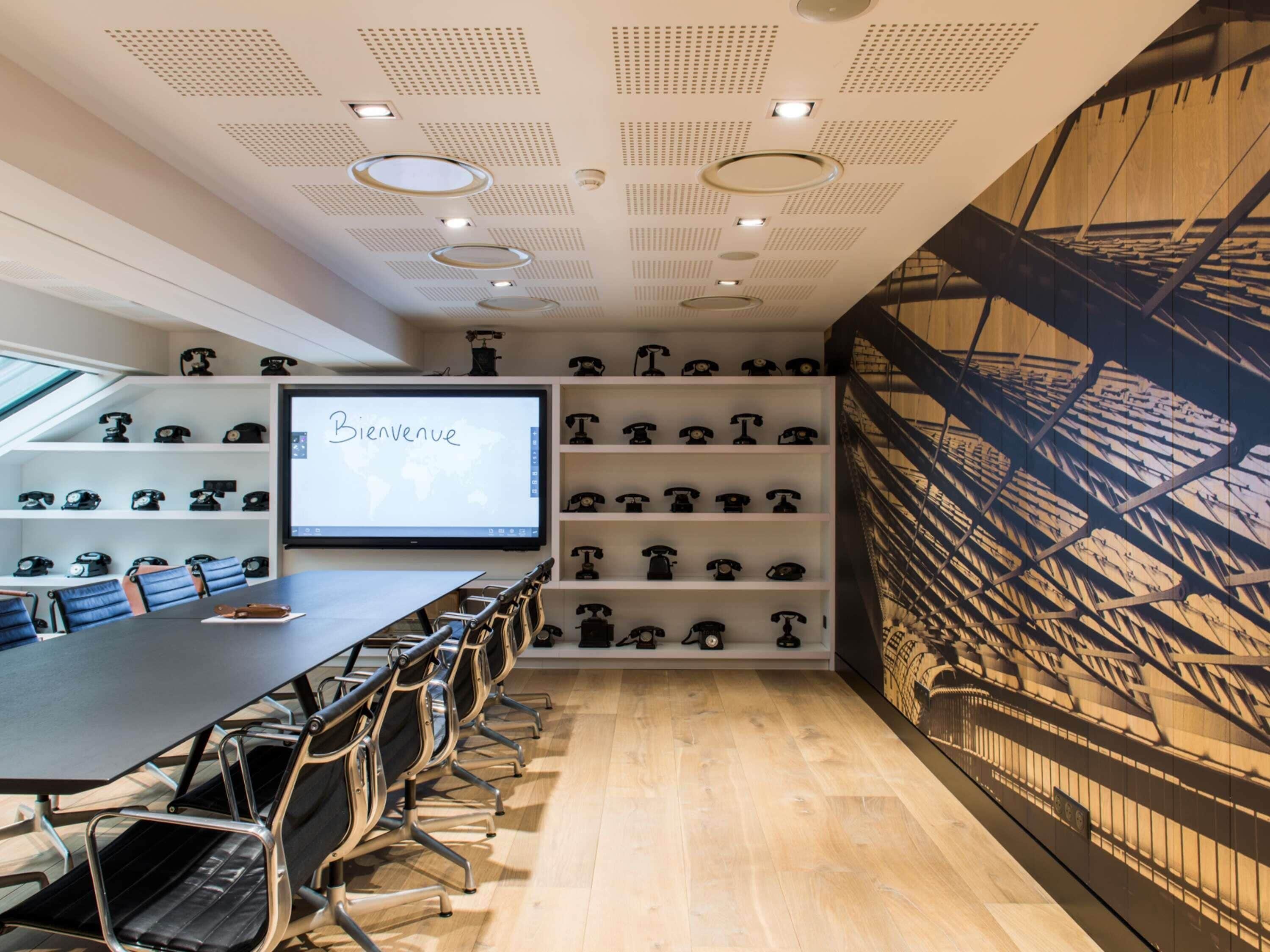 A conference room with a large screen displaying 'Bienvenue,' a wall of vintage telephones, and a ceiling with recessed lighting.